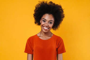 Neşeli Afrikalı Amerikalı kız gülümsüyor ve sarı arka planda izole edilmiş kameraya bakıyor.
