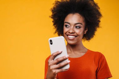 Sarı arka planda izole edilmiş cep telefonu kullanırken gülümseyen Afrikalı Amerikalı kız.