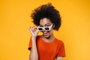 Güneş gözlüklü neşeli Afrikalı Amerikalı kız sarı arka planda yalnız başına gülümsüyor ve yan tarafa bakıyor.