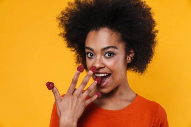 Sarı arka planda izole edilmiş ahududu yerken gülümseyen Afrikalı Amerikalı kız.