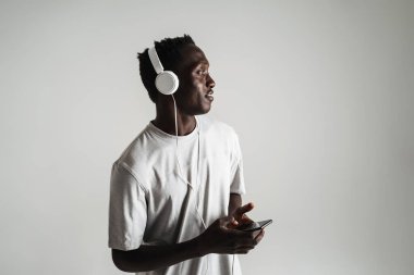 Gri arka planda izole edilmiş kulaklıkla müzik dinleyen, günlük kıyafetli genç Afrikalı bir adam.
