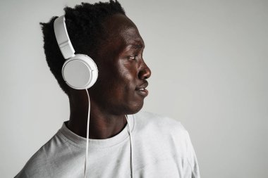Gri arka planda izole edilmiş kulaklıkla müzik dinleyen, günlük kıyafetli genç Afrikalı bir adam.