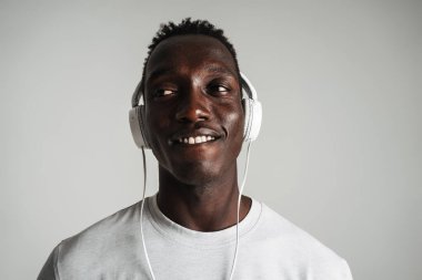 Gri arka planda izole edilmiş kulaklıklarla müzik dinleyen sıradan giyinmiş mutlu Afrikalı genç bir adam.