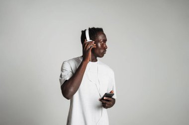 Gri arka planda izole edilmiş kulaklıkla müzik dinleyen sıradan giyinmiş Afrikalı genç bir adam.