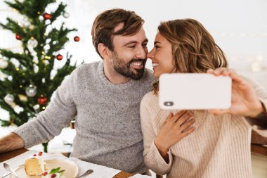 Çekici, neşeli bir çift cep telefonuyla fotoğraf çekiyor ve rahat bir odada Noel yemeği yiyor.