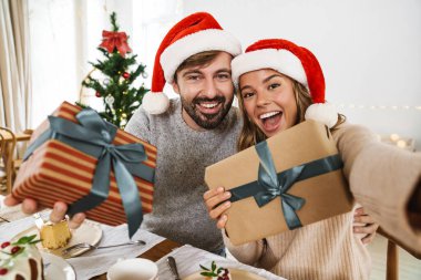 Güzel, heyecanlı bir çift hediye kutularıyla selfie çekerken rahat bir odada Noel yemeği yiyor.