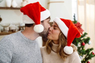 Noel Baba şapkalı mutlu çift evde Noel ağacının yanında öpüşüyor.