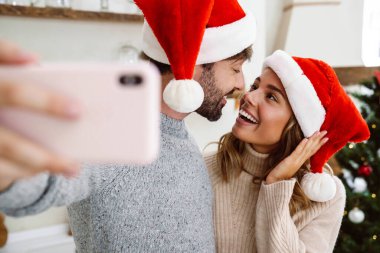 Yılbaşı kıyafeti giymiş genç bir çiftin parlak bir mutfakta cep telefonuyla selfie çekerken görüntüsü.