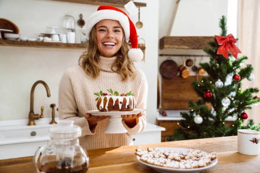 Noel Baba şapkalı mutlu kadın rahat bir mutfakta Noel pastasıyla poz veriyor.