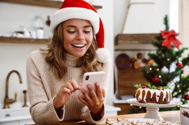Noel Baba şapkalı mutlu bir kadın. Mutfakta cep telefonu kullanırken gülümsüyor.