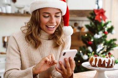 Noel Baba şapkalı mutlu bir kadın. Mutfakta cep telefonu kullanırken gülümsüyor.