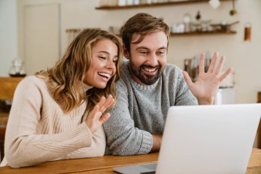 Güzel mutlu çift el sallıyor ve rahat bir mutfakta otururken dizüstü bilgisayar kullanıyor.