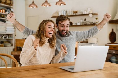 Güzel heyecanlı çift, rahat bir mutfakta dizüstü bilgisayar kullanırken güzel bir jest yapıyor.