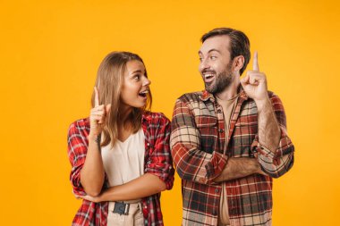 Heyecanlı güzel bir çift, sarı arka planda, parmaklarını yukarı kaldırarak işaret ediyorlar.