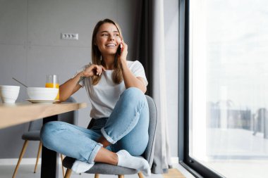 Güzel, neşeli kız evde kahvaltı ederken cep telefonuyla konuşuyor.