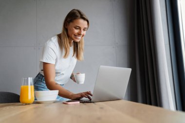 Ev mutfağında kahve içerken cep telefonu ve dizüstü bilgisayar kullanan güzel mutlu bir kız.