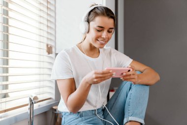 Ev mutfağında otururken cep telefonu kullanan kulaklıklı güzel bir kız.