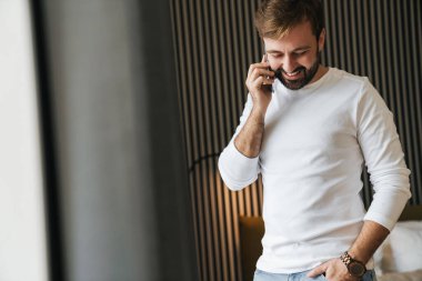 Çekici gülümseyen genç adam yatak odasında dururken cep telefonu kullanıyor.