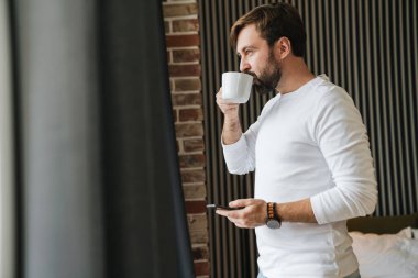 Yatak odasında kahve içerken cep telefonu kullanan kendine güvenen çekici genç adam.