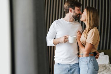 Mutlu genç çift yatak odasında ayakta kahve içerken kucaklaşıyorlar.