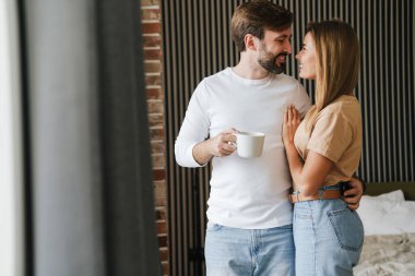 Mutlu genç çift yatak odasında ayakta kahve içerken kucaklaşıyorlar.
