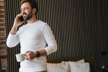 Çekici gülümseyen genç adam yatak odasında durup kahve içerken cep telefonu kullanıyor.