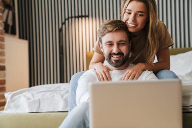 Laptop bilgisayarıyla arkadaşlarıyla video görüşmesinde, yatak odasında oturan mutlu, genç ve çekici çift.