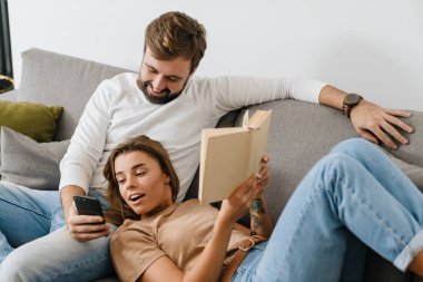 Genç çift kanepede birlikte rahatlıyor, kitap okuyor ve cep telefonu kullanıyor.