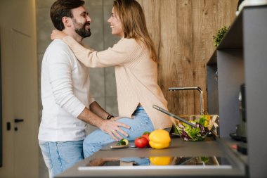 Gülümseyen genç çift mutfakta salata pişiriyor, sarılıyorlar.