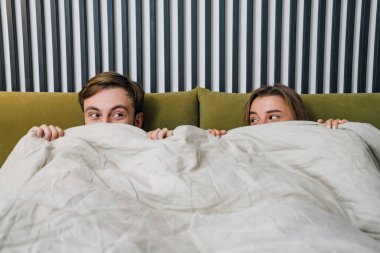 Mutlu genç çift yatakta yatarken battaniyeden dışarı bakıyor.