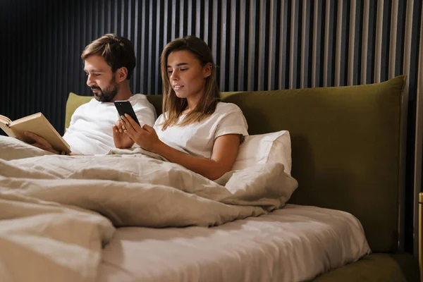 Glückliches Junges Paar Liest Buch Und Benutzt Handy Bett — Stockfoto