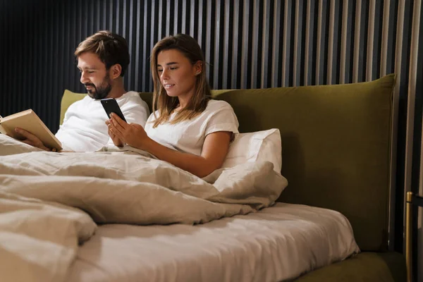 Glückliches Junges Paar Liest Buch Und Benutzt Handy Bett — Stockfoto