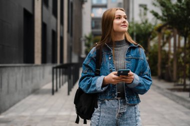 Kot pantolon giyen güzel genç bir kadın sokakta yürüyor, cep telefonu kullanıyor.