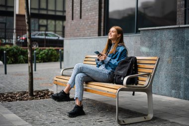 Şehir caddesinde bankta otururken cep telefonu kullanan güzel, neşeli öğrenci kız.