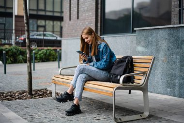 Kot pantolon giyen güzel genç bir kadın bir bankta oturuyor, cep telefonu kullanıyor.