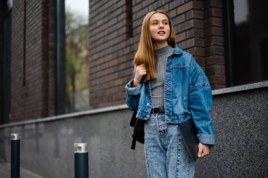 Kot pantolon giyen güzel genç bir kadın sokakta yürüyor, dizüstü bilgisayar taşıyor.