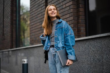 Güzel mutlu öğrenci kız gülümseyerek ve dizüstü bilgisayarını tutarak şehir caddesinde duruyor.