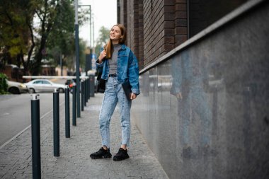 Güzel mutlu öğrenci kız gülümseyerek ve dizüstü bilgisayarını tutarak şehir caddesinde duruyor.