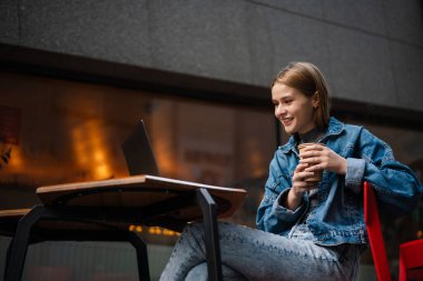 Genç, güzel bir kız bir kafede oturuyor ve elinde kahve fincanı tutarak bilgisayarla çalışıyor.