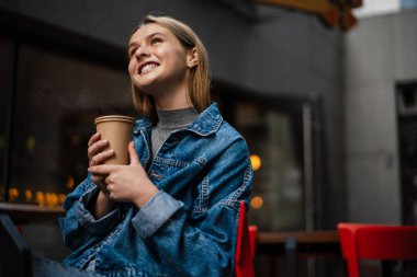 Güzel, neşeli bir öğrenci kız sokak kafesinde otururken kahve içiyor.