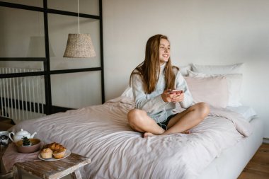 Güzel, gülümseyen, güzel bir kız. Evde yatakta otururken cep telefonu ve kulaklık kullanıyor.