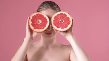 Memnun genç bir kadın, elinde greyfurtla stüdyoda pembe bir arka planda poz veriyor.