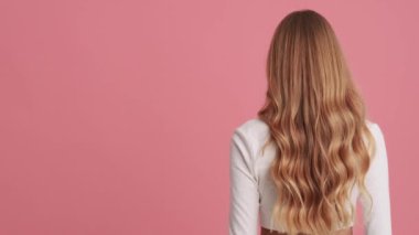 Şok olmuş genç sarışın bir kadın kameraya dönüyor stüdyodaki pembe bir duvarın üzerinde tek başına duruyor.