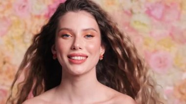 Mutlu genç bir kadının yakın çekim görüntüsü kameraya poz verirken rüzgarda dalgalanan saçlar stüdyodaki çiçekli bir arka planda izole bir şekilde duruyor.