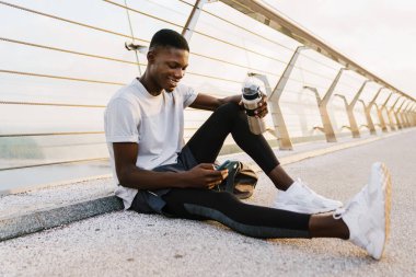 Yakışıklı Afrikalı sporcu cep telefonu kullanıyor, elinde su şişesi şehir köprüsünde oturuyor.