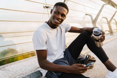 Yakışıklı Afrikalı sporcu cep telefonu kullanıyor, elinde su şişesi şehir köprüsünde oturuyor.