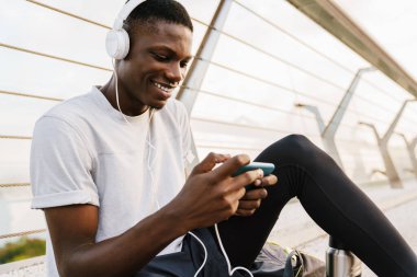 Rahatlamış gülümseyen Afrikalı sporcu köprüde oturuyor, kulaklık takıyor, cep telefonu kullanıyor.