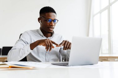 Resmi kıyafetler ve gözlük takan çekici, koyu tenli bir iş adamı müşterisiyle iş görüşmelerini dizüstü bilgisayarda, kulaklıkla yapıyor.