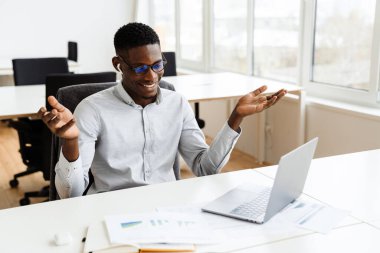 Resmi kıyafetler ve gözlük takan çekici, koyu tenli bir iş adamı müşterisiyle iş görüşmelerini dizüstü bilgisayarda, kulaklıkla yapıyor.