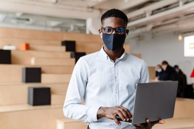 Tıbbi maske takan kendinden emin Afrikalı bir öğrenci sınıfta dizüstü bilgisayarını tutuyor.